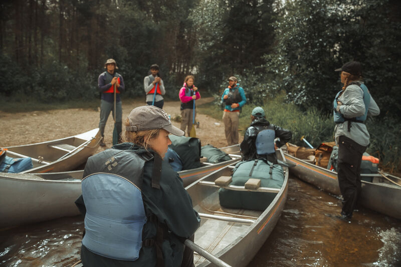 A group of seven people are gathered near several canoes, seemingly preparing for a water adventure. They are dressed in outdoor attire, with some wearing life jackets, suggesting they are about to embark on a canoeing trip. The canoes are lined up on the edge of a body of water, with a forest visible in the background, indicating a natural setting for their activity.
